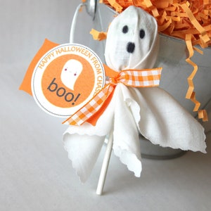 Pode incluir: Uma pirulito de fantasma de tecido branco com olhos pretos e uma fita xadrez laranja e branca. A pirulito est&aacute; presa a um pau branco e tem uma etiqueta que diz "Feliz Halloween do Chase! Boo!"