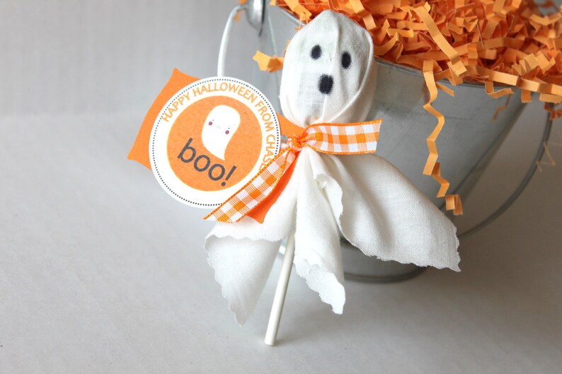 Pode incluir: Uma pirulito de fantasma de tecido branco com olhos pretos e uma fita xadrez laranja e branca. A pirulito est&aacute; presa a um pau branco e tem uma etiqueta que diz "Feliz Halloween do Chase! Boo!"