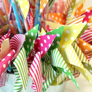 May include: A close-up of a colorful bouquet of paper pinwheels in a white bucket. The pinwheels are made of paper with different patterns, including polka dots, stripes, and swirls. The pinwheels are in various colors, including red, green, yellow, and orange.