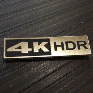 Könnte beinhalten: Ein rechteckiges Metallabzeichen mit dem Text "4K HDR". Die linke Seite des Abzeichens hat die Zahl "4K" in einem schwarzen Rechteck, während die rechte Seite "HDR" in Gold hat.