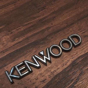 Könnte beinhalten: Nahaufnahme eines schwarzen und silbernen Kenwood-Logos auf einer Holzoberfläche. Die Buchstaben sind schwarz umrandet mit silbernem Innenraum, wodurch ein 3D-Effekt entsteht. Der Holzmaserungshintergrund ist dunkelbraun.