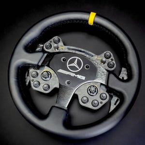 Può includere: Un volante AMG nero con una striscia gialla nella parte superiore. Il volante presenta diversi pulsanti e quadranti, e il logo Mercedes-Benz è ben visibile al centro. Il volante è in pelle.