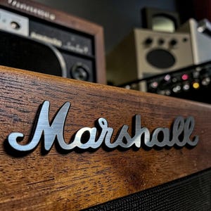 Può includere: Primo piano di un amplificatore in legno con il logo "Marshall" argentato. L'amplificatore ha una finitura in legno scuro e una griglia per altoparlanti nera. Altre apparecchiature audio sono visibili sullo sfondo.