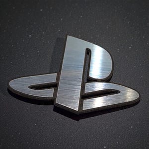 Può includere: Logo PlayStation argentato con texture in metallo spazzolato. L'iconico simbolo, composto da una 'P' stilizzata e una linea orizzontale, è associato ai videogiochi. Il logo è posto su uno sfondo scuro.