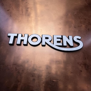 Può includere: Logo Thorens argento e nero, con effetto metallo spazzolato, su sfondo color rame. Il logo, in carattere sans-serif, presenta una curva stilizzata sotto la parola.