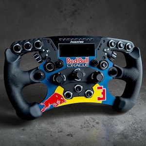May include: A black and blue racing steering wheel controller featuring the Red Bull Oracle logo, a central digital display, multiple buttons, rotary dials, and textured black grips. The design includes a red bull graphic and the number 3.