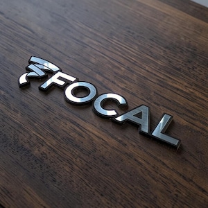 Op de afbeelding: Zilverkleurig metalen "FOCAL" logo op een donkerbruin houten oppervlak. Het logo heeft een geborsteld metalen uiterlijk met een zwarte omtrek. Het woord "FOCAL" is in een schreefloos lettertype.