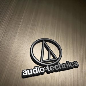 Puede incluir: Logotipo negro y plateado de Audio-Technica, con un triángulo dentro de un círculo y el nombre de la marca debajo. El logotipo está sobre un fondo dorado metálico texturizado.