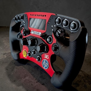 Könnte beinhalten: Ein rot-schwarzes Rennlenkrad im Formel-1-Stil mit zentralem Digitaldisplay, mehreren Drehknöpfen und Drucktasten. Das Gerät zeigt auf der Vorderseite das Scuderia Ferrari-Logo, die Nummer 16, ein Shell-Emblem sowie ein HP-Logo.