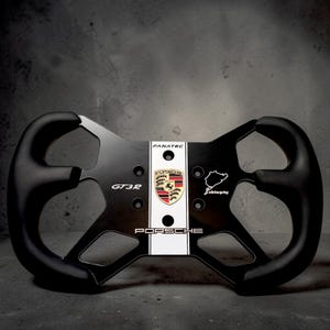 Puede incluir: Volante de carreras negro y plateado con el logotipo de Porsche, texto GT3R y contorno de Nürburgring. El volante tiene una franja blanca en el centro con el logotipo de Fanatec.