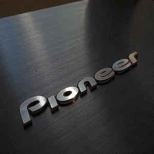 Könnte beinhalten: Nahaufnahme einer schwarzen, strukturierten Oberfläche mit dem Wort "Pioneer" in silbernen Buchstaben. Das Wort ist in einer stilisierten Schriftart gehalten. Die Oberfläche hat einen gebürsteten Metalleffekt, mit Lichtreflexen.