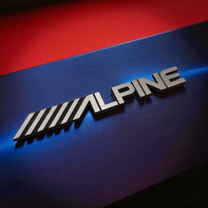 May include: A close-up shot of the silver "ALPINE" logo on a blue surface with a red upper section. The logo has a distinctive design with diagonal stripes. The background has a brushed metal texture.
