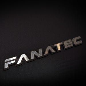May include: A silver and black "FANATEC" emblem, with a brushed metal finish, is displayed against a dark, textured background. The letters are slightly raised, creating a three-dimensional effect. The emblem is angled.