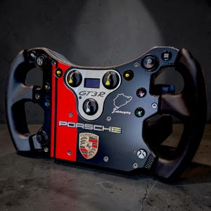 Peut inclure: Un volant de course Porsche GT3R noir et rouge avec l'écusson Porsche au centre, le tracé du Nürburgring et divers boutons marqués N, P, X, Y et des icônes de menu. L'appareil présente une texture en fibre de carbone sur les côtés.