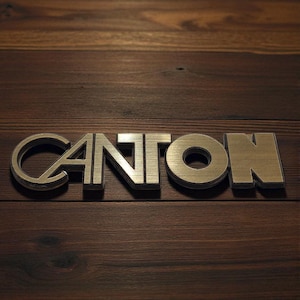Op de afbeelding: Het woord "CANTON" in metallic letters wordt weergegeven op een donkerbruin houten oppervlak. De letters zijn dik en driedimensionaal, met een geborsteld metaaleffect, waardoor subtiele schaduwen ontstaan.