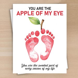 May include: A white greeting card featuring a watercolor illustration of two red baby footprints shaped like an apple with a green leaf and brown stem. The text reads YOU ARE THE APPLE OF MY EYE and You are the sweetest part of every season of my life.
