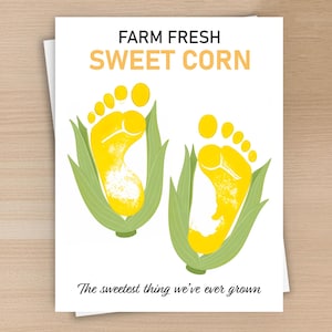 May include: A white greeting card featuring the text FARM FRESH SWEET CORN at the top and The sweetest thing we’ve ever grown at the bottom. The centre displays two yellow baby footprint illustrations, each partially wrapped in green corn husks.