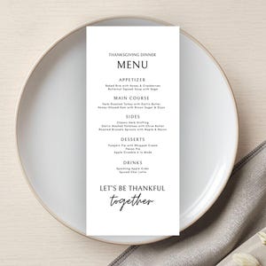 Könnte beinhalten: Eine Speisekarte für das Thanksgiving-Dinner, gedruckt auf einer weißen Karte, die auf einem weißen Teller liegt. Die Speisekarte umfasst Vorspeisen, Hauptgerichte, Beilagen, Desserts und Getränke. Auf der Karte steht "Let's be thankful together".