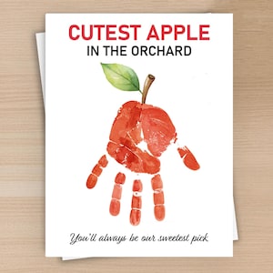 May include: A white greeting card featuring a red handprint shaped like an apple with a green leaf and brown stem. The text reads CUTEST APPLE IN THE ORCHARD at the top and You'll always be our sweetest pick in black cursive script at the bottom.