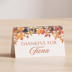 May include: A folded white place card featuring a top border of blue and white plaid fabric, orange and yellow autumn maple leaves, and glowing string lights. The text THANKFUL FOR Fiona is printed in the center in a dark orange serif font.