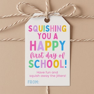 May include: A white rectangular gift tag with rounded top corners, tied with tan twine. The text reads: SQUISHING YOU A HAPPY first day of SCHOOL! Have fun and squish away the jitters! FROM: The lettering uses various colors including pink, blue, yellow, and green.