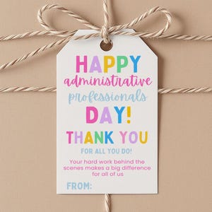 Puede incluir: Un regalo envuelto en papel beige, atado con hilo y una etiqueta blanca. La etiqueta dice "HAPPY administrative professionals DAY! THANK YOU FOR ALL YOU DO!" en texto colorido. La etiqueta también incluye el texto "Your hard work behind the scenes makes a big difference for all of us" y una línea "FROM:"