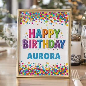 May include: A framed birthday sign with the words "HAPPY BIRTHDAY AURORA" in colorful letters. The sign is decorated with confetti and stars in various colors, set against a white background.