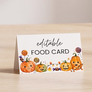 May include: A white folded food card featuring a watercolor illustration of three smiling pumpkins, colorful lollipops, candy corn, and a string of orange lights. The text "editable" is written in black script above the text "FOOD CARD" in black.