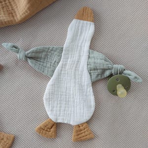 May include: A white, duck-shaped baby comforter with a brown beak and feet, and light green wings. A green pacifier is attached to one wing. The comforter is made of soft, textured fabric.