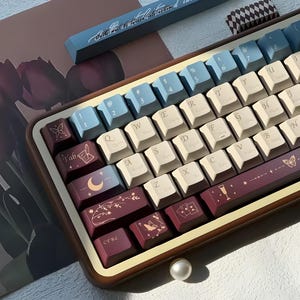May include: A custom mechanical keyboard with a wooden frame and keycaps in cream, burgundy, and blue. The keycaps feature symbols and text, including the word "Tab" and the numbers 1-8. Decorative butterfly and moon designs are also present.