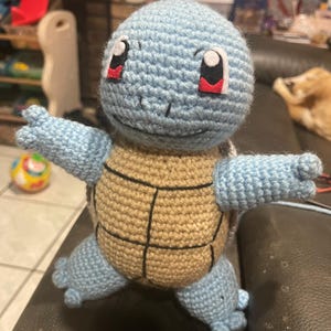 May include: A handmade crochet Squirtle plushie. The plushie features a light blue head, arms, and feet, with a tan shell. It has black eyes with white highlights and red accents. The toy is sitting on a dark surface.