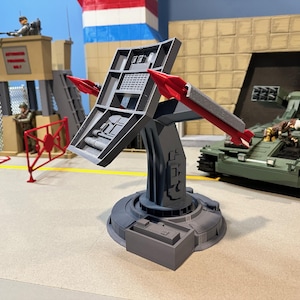 May include: A gray missile launcher with two red missiles, part of a military diorama. The scene includes a green tank, a watchtower, and figures of soldiers. The launcher is on a gray base.