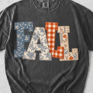 May include: A charcoal gray short-sleeve t-shirt featuring the word FALL in large block letters. The letters are filled with patterns including blue floral, cream floral, and orange gingham check, with small orange pumpkin illustrations.
