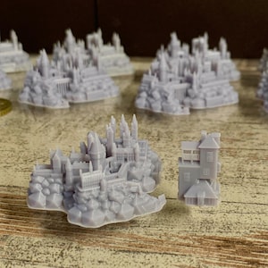 May include: A collection of 3D printed miniature buildings, including a large castle and a smaller, two-storey house. The buildings are a light grey colour and are set on a wooden surface.