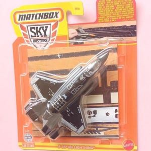 Op de afbeelding: Matchbox Sky Busters F-35 (B) Lightning speelgoedvliegtuig in een blisterverpakking. Het zwart-zilveren speelgoedvliegtuig heeft gedetailleerde kenmerken. De verpakking is oranje en geel met het Matchbox-logo en de tekst "Sky Busters".