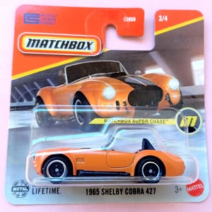 Op de afbeelding: Een Matchbox 1965 Shelby Cobra 427 gegoten speelgoedauto in oranje met zwarte wielen, verpakt in een blisterkaart met een oranje en zwarte afbeelding van het voertuig, het Matchbox-logo en de tekst Matchbox Super Chase en 3/4.