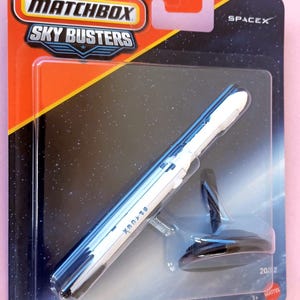 May include: A Matchbox Sky Busters SpaceX Falcon Heavy die-cast rocket toy in its original blister pack packaging. The white rocket features the SpaceX logo and is displayed with a black plastic stand inside the clear plastic bubble casing.