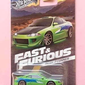 Può includere: Un'auto giocattolo in metallo pressofuso verde del 1995 Mitsubishi Eclipse in una confezione blister di plastica. L'auto ha una grafica blu e bianca sul lato e le parole "Fast & Furious Brian O'Conner" sulla confezione.
