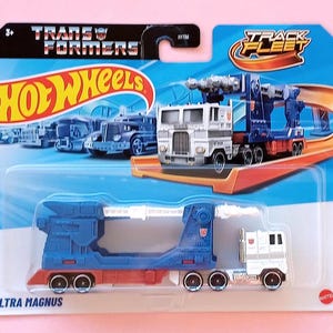 Może przedstawiać: Niebiesko-biały plastikowy samochód ciężarowy z czerwonym paskiem. Ciężarówka jest zabawką Hot Wheels Transformers Track Fleet Ultra Magnus. Ciężarówka znajduje się na niebiesko-białym tle z napisami "Transformers" i "Hot Wheels".