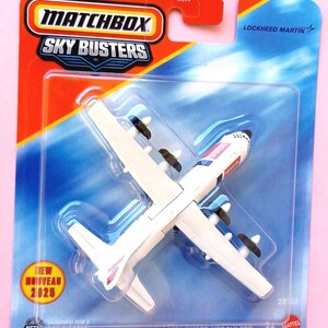 May include: A white Matchbox Sky Busters Lockheed C-130J Super Hercules toy aeroplane in its original packaging. The plane features black propellers and red Rescue text on the fuselage. The card includes the Matchbox logo and Lockheed Martin branding.