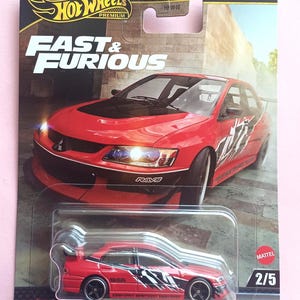 Może przedstawiać: Model odlewu Hot Wheels Premium czerwonego Mitsubishi Lancer Evolution IX z serii Fast & Furious. Samochód ma czarno-białą grafikę, czarne koła i napis "RAYS". Opakowanie zawiera logo Hot Wheels i numer 2/5.
