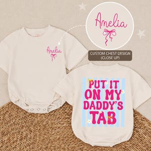 May include: Off-white baby bodysuit with short sleeves. The front features the name "Amelia" in pink script with a bow graphic. The back has the text "PUT IT ON MY DADDY'S TAB" in pink and blue, with floral accents. The bodysuit is displayed on a woven surface.