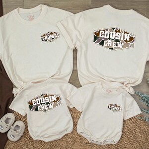May include: Two off-white t-shirts and two off-white baby bodysuits. One t-shirt features a small camo patch with the name Thomas, the other a large camo patch with the text Cousin Crew. The bodysuits mirror these designs with the same camo patterns.
