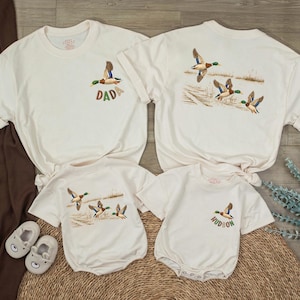 May include: Off-white t-shirts and baby rompers with duck embroidery. One shirt says "DAD" and another has a duck scene on the back. A baby romper says "HUDSON". The clothing is on a woven mat.