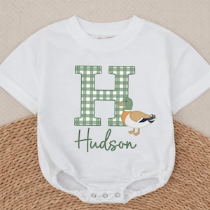 May include: A white short-sleeved bodysuit featuring a large letter H in a green and white gingham pattern. Next to the letter is a graphic of a mallard duck with a green head, brown body, and blue wing detail. The name Hudson is printed below in green.
