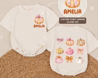 May include: Two cream-colored short-sleeve baby bodysuits. One features a small pumpkin with a pink bow and the name AMELIA. The other displays a grid of nine illustrations including pumpkins, bows, and plaid patterns in orange, pink, and white.