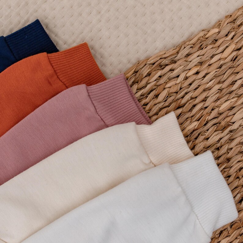 Op de afbeelding: Close-up van de manchetten van verschillende sweatshirts in marineblauw, oranje, roze, cr&egrave;me en wit. De geribbelde manchetten zijn diagonaal over een gevlochten mand geplaatst. De afbeelding toont de verscheidenheid aan kleuren.