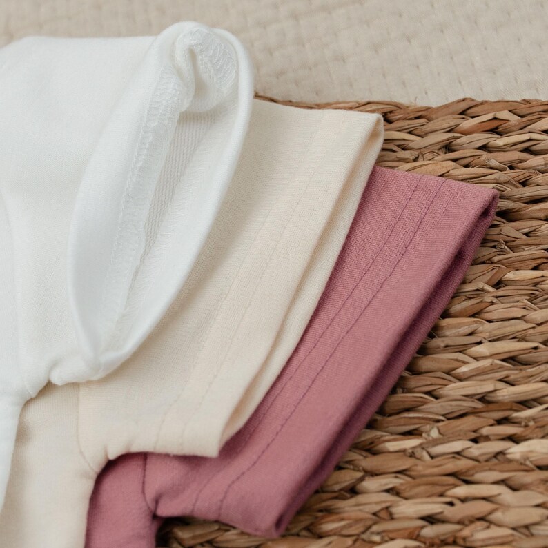 Op de afbeelding: Close-up van drie opgevouwen t-shirts in wit, cr&egrave;me en stoffig roze. Het witte shirt staat bovenaan, gevolgd door cr&egrave;me en dan stoffig roze. De shirts zijn gemaakt van zachte stof en zijn netjes opgevouwen. De shirts liggen op een geweven oppervlak.