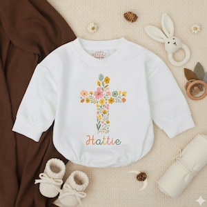 Op de afbeelding: Witte babyromper met een bloemenkruisontwerp. Het kruis is versierd met kleurrijke bloemen en groen. De naam "Hattie" is in een regenbooglettertype eronder gedrukt. De romper heeft lange mouwen en een ronde halslijn.