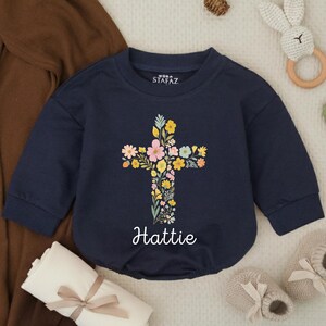 Op de afbeelding: Marineblauwe babyromper met een bloemenkruisontwerp. Het kruis bestaat uit kleurrijke bloemen in roze, geel en blauw. De naam "Hattie" staat in sierlijke letters eronder. De romper heeft lange mouwen en een ronde halslijn.